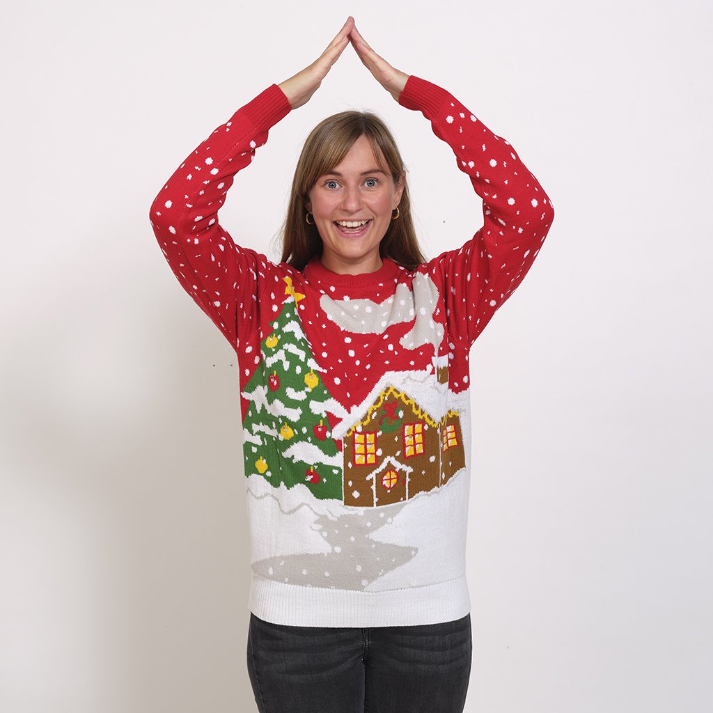 Une femme portant un pull de Noël rouge avec une maison enneigée et un arbre de Noël illuminé.